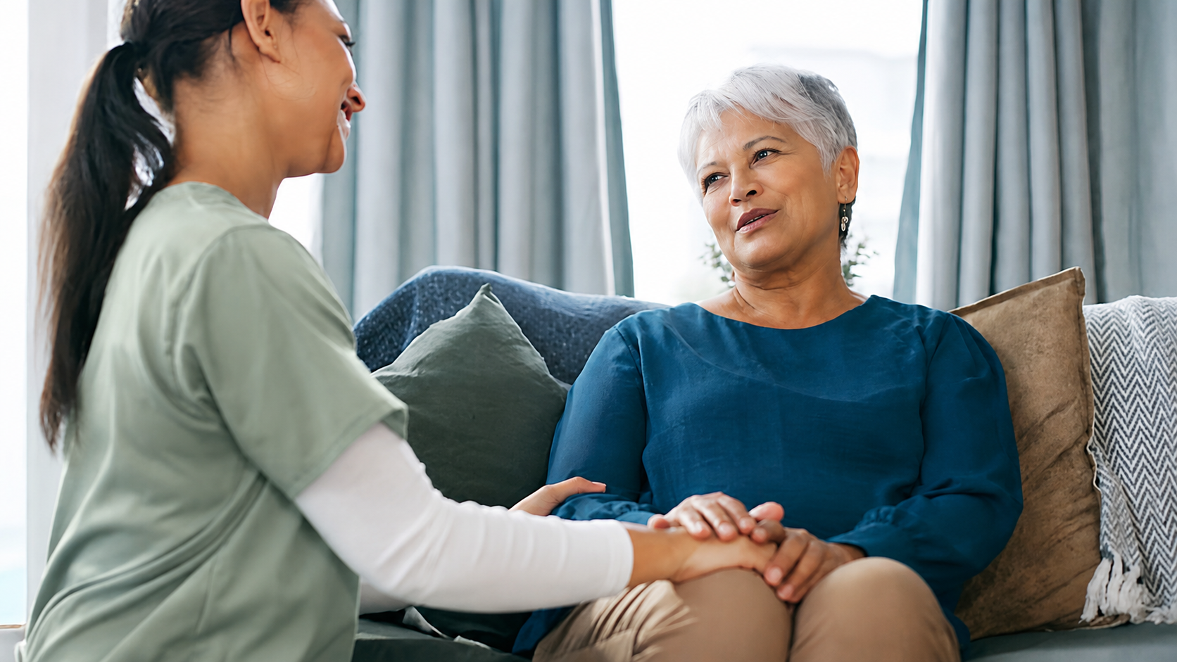 A caregiver holding a senior client's hands while they talk on a sunlit living room sofa